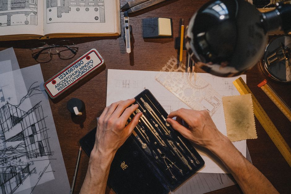 Overhead view of an architect's hands sketching interior floor plans with drafting tools on a wooden desk